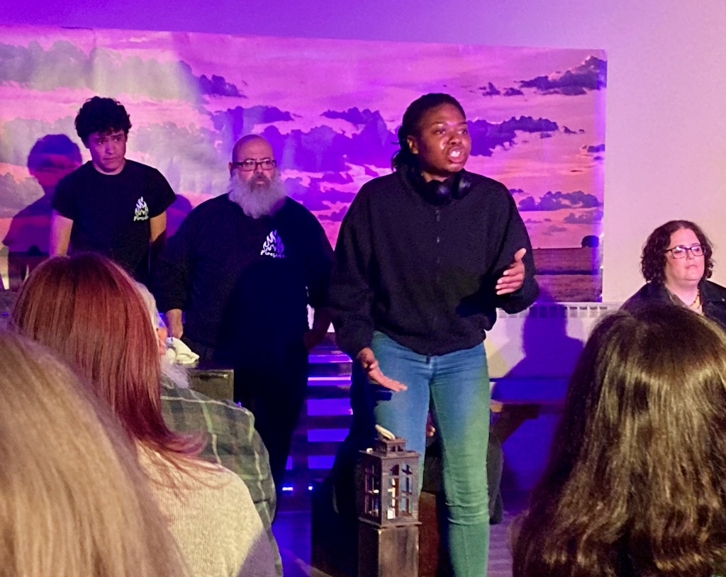 A Black actor is emphatically speaking, with the ensemble behind them and the audience in front.